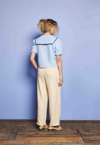 Kvinne med krøllete hår står vendt mot en lavendelvegg, iført en lyseblå topp med sjømannskrage og kremfargede vide bukser med sandaler.