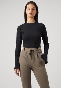 Νεαρή γυναίκα που φοράει μαύρο ριμπ μπλουζάκι με μακριά μανίκια και μπεζ ψηλόμεσα παντελόνια με κορδόνι, στέκεται μπροστά σε απλό φόντο.