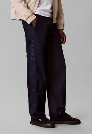 Person trägt schwarze Calvin Klein Hose mit Seitlichem Logo, beige Jacke, weißes Hemd und schwarze Turnschuhe, steht vor grauem Hintergrund.