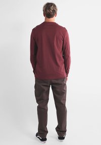 Chemise à manches longues bordeaux avec un col rond, associée à un pantalon marron. La tenue est complétée par des baskets noires.