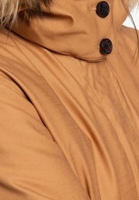 Chaqueta marrón con un cuello alto, que presenta dos botones negros y detalles de costura visibles. Textura suave y brillo sutil.