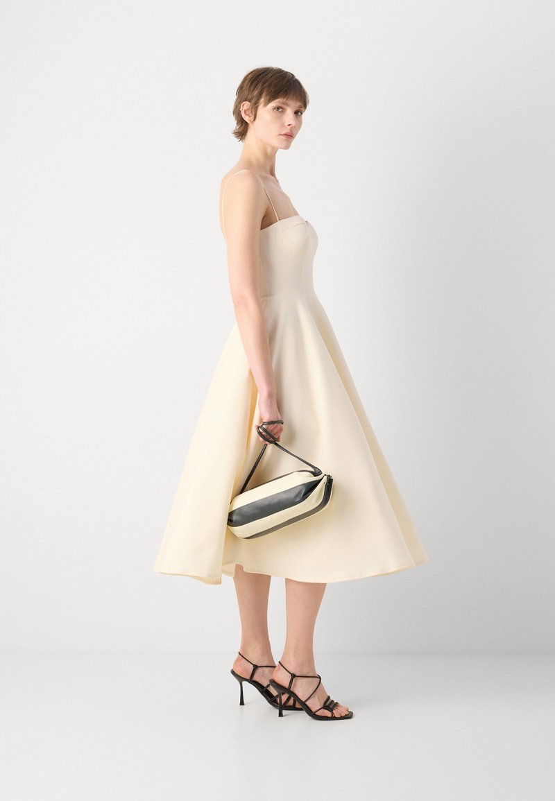 Cremefarbenes Midikleid mit dünnen Trägern, schwingendem Rock und strukturiertem Stoff. Eine schwarz-weiße Handtasche und schwarz-weiße Riemchen-Pumps runden den Look ab.