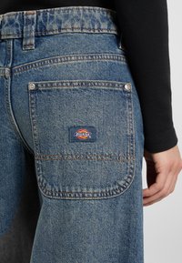 Close-up of blue denim jeans back pocket with Dickies logo patch, worn by person in black long-sleeve shirt.