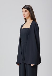 Kimono noir à manches longues, ouvert, porté sur un top sans manches ajusté. Tissu lisse, design simple et silhouette décontractée avec des manches larges.