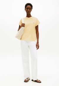 T-shirt gialla a maniche corte con un piccolo logo, pantaloni bianchi a gamba larga e infradito neri. Il modello porta una borsa beige.