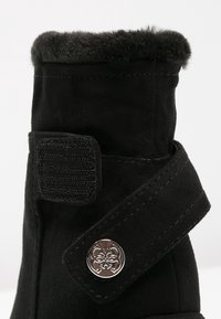 Botte en suède noire avec un col en fausse fourrure, une sangle en velcro ajustable et un bouton en métal décoratif sur le côté.
