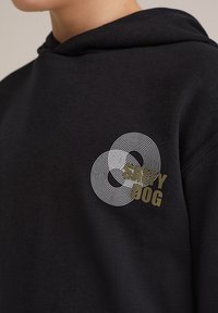 Melnā kapuce ar baltu apaļu līnijas dizainu un tekstu "SALTY DOG" olīvu zaļā krāsā. Izgatavota no mīkstas, teksturētas auduma ar kangaroo kabatu.