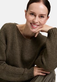 Eine Frau mit braunen Haaren lächelt, während sie ihr Kinn auf ihre Hand stützt. Sie trägt einen grob gestrickten dunkeloliven Pullover vor einem einfarbigen Hintergrund.