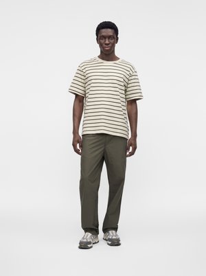 Bărbat stând în picioare, purtând un tricou în dungi bej și verde, pantaloni verde măsliniu și adidași gri, pe un fundal alb simplu.