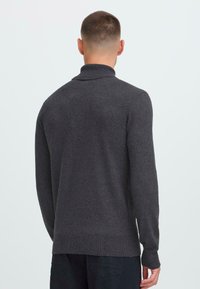 Uomo con capelli corti che indossa un maglione a maniche lunghe grigio scuro e pantaloni neri, in piedi con le spalle rivolte verso di noi su uno sfondo chiaro uniforme.