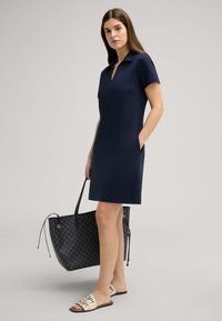 Navyblauw polo jurk met korte mouwen, met een v-hals en zijzakken. Vergezeld door een gestreepte tote bag en beige sandalen.