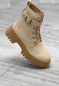 Beige semsket ankle boot med rund tå, snøring foran, justerbar stropp og en tykk gummisåle med mønster.