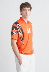 Orange tröja med svartvita grafiska mönster, sportig krage och ett vitt nummer "94" på framsidan. Bärs med ljusfärgade byxor.