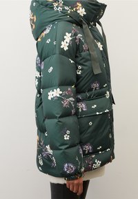 Mörkgrön vadderad jacka med flerfärgad blommönster, stor huva, dragsko, elastiska ärmar och framfickor visad på en person.