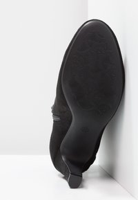 Botín de ante negro con tacón delgado, que presenta una suela de goma texturizada con un patrón floral y un detalle de cremallera lateral. Talla marcada como 37.