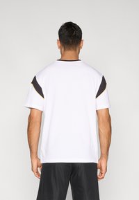 Άσπρο μπλουζάκι με μαύρες και κίτρινες πινελιές στα μανίκια, κοντά μανίκια, στρογγυλή λαιμόκοψη και άνετη εφαρμογή. Η πίσω όψη παρουσιάζει απλό σχέδιο.