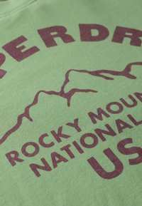 Groene katoenen sweatshirt met donkerpaarse tekst en een bergsilhouet, met de tekst "ROCKY MOUNTAIN NATIONAL PARK, VS." Zachte textuur.