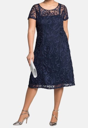Frau trägt knie-langes marineblaues Kleid mit durchsichtigem besticktem Überzug, hält silberne Clutch und trägt silberne Riemchen-High-Heels.