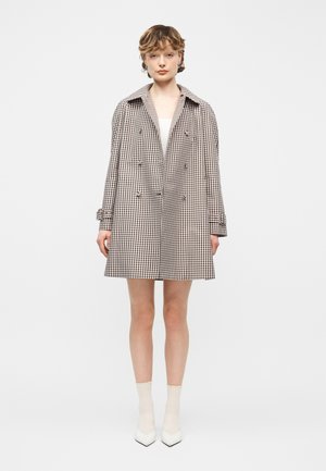 Giovane in piedi, indossa un cappotto a quadri marrone e bianco fino al ginocchio, maglietta bianca, calzini bianchi e scarpe bianche con la punta a punta, sfondo neutro.