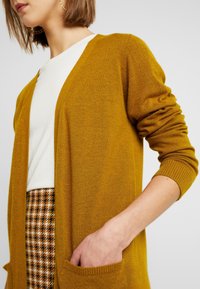 Senfgelber Cardigan mit langen Ärmeln, offenem Vorderteil, gerippten Bündchen und zwei Taschen. Über einem weißen Hemd und einem gemusterten Rock getragen. Weicher Stoff.