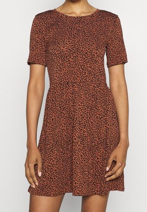 Femme portant une robe marron à manches courtes imprimée léopard avec une taille froncée, debout devant un fond clair uni.