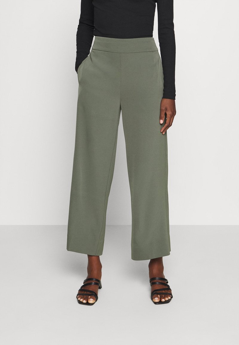Personne portant un pantalon large taille haute vert olive, un haut noir à manches longues et des sandales noires à brides, debout sur un fond uni.