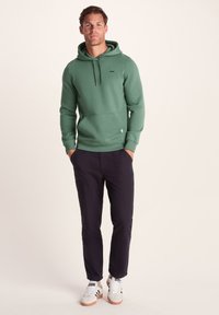Green hoodie with a drawstring, front pocket, and logo patch, paired with dark pants and white sneakers featuring black stripes.