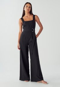 Žena stojící s rukou v kapse, oblečená v černém bezrukávovém širokém overal s opaskem a béžových sandálech na jednobarevném pozadí.