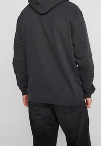 Henkilö, joka on pukeutunut tummanharmaaseen huppariin ja mustiin farkkuihin, kääntynyt poispäin vaaleanharmaata taustaa vasten.