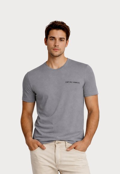 Νέος άνδρας στέκεται με το πρόσωπο προς τα εμπρός, φορώντας εφαρμοστό γκρι t-shirt Emporio Armani και μπεζ παντελόνι, με τα χέρια στις τσέπες.