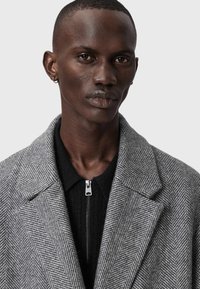 Pelēks zigzagmājas mētelis ar plašiem atlociem, uzvilkts virs melna ar rāvējslēdzi adīta topa. Mētelis ir izgatavots no teksturēta auduma un tam ir pulēts sudraba rāvējslēdzis.