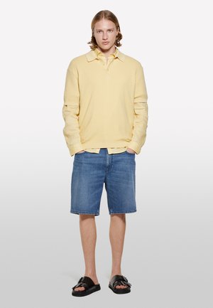 Polo shirt en coton jaune avec un motif texturé, porté par-dessus une chemise à boutons plus claire ; associé à un short en denim bleu et des sandales noires.