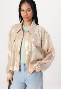 Sequin jacket in beige, featuring a button-up front, two chest pockets, and a relaxed fit. Worn over a light green top and blue jeans.