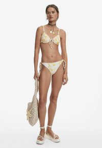 Witte bikini met geel bloemenpatroon, voorzien van verstelbare strikjes. Vergezeld van een gevlochten tas en platform sandalen. Soepele stoftextuur.