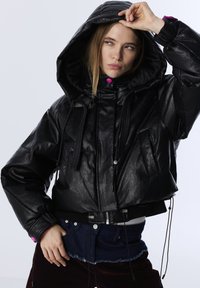 Vrouw in een zwarte leren jack met capuchon en roze knopen die de capuchon optilt, gecombineerd met donkerblauwe denim en bordeauxrode broeken.