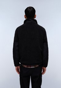 Giacca in pile nera con colletto alto, tessuto strutturato e maniche imbottite nere. Indossata sopra una camicia a quadri, con pantaloni cargo visibili.