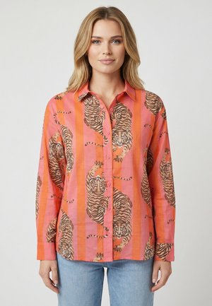 Woman wearing a pink and orange striped button-up shirt with tiger prints, paired with light blue jeans, standing against a plain background.