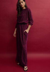 Robe fuchsia à manches longues avec des rayures verticales, dotée d'une taille cintrée et de poches latérales. Associée à une ceinture noire et des tongs.