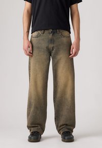 Χαλαρά, ξεθωριασμένα τζιν σε ανοιχτό καφέ απόχρωση με πιο σκούρες λεπτομέρειες. Σχεδίαση με πέντε τσέπες, ορατές ραφές και κανονική γραμμή στα πόδια.