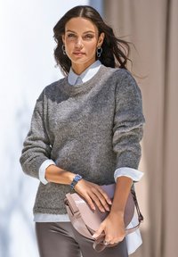 Grau melierter Strickpullover mit Rundhalsausschnitt, über einem weißen Hemd mit Kragen getragen. Das Model hält eine hellrosa Umhängetasche.