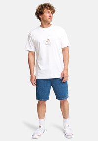 White cotton t-shirt with a printed logo in brown, paired with blue denim shorts. Model wears white sneakers and white crew socks.