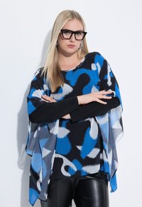 Top a fantasia nera e blu con una vestibilità ampia, maniche drappeggiate, indossato sopra una maglietta nera a maniche lunghe, abbinato a pantaloni di pelle nera.
