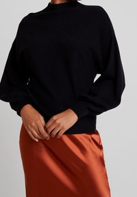 Zwarte gebreide trui met hoge kraag en ballonmouwen, gecombineerd met een oranje satijnen rok. Zachte textuur, getailleerd ontwerp en zichtbare manchetdetails.