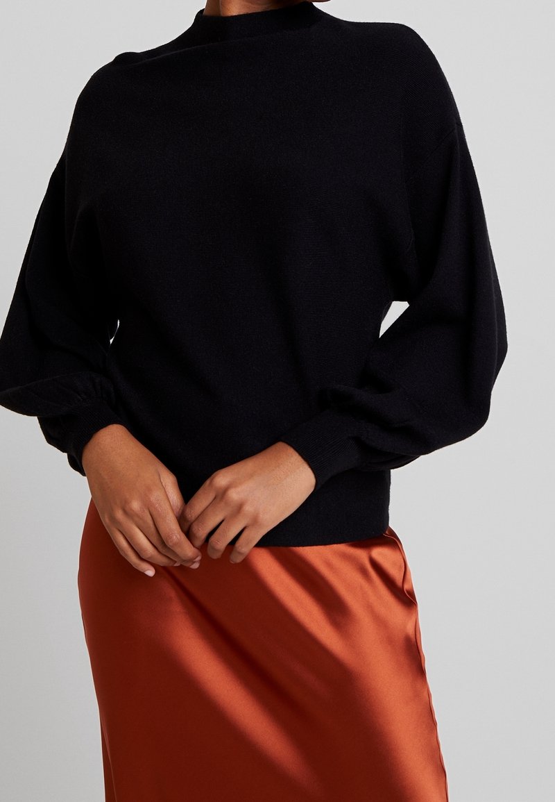 Zwarte gebreide trui met hoge kraag en ballonmouwen, gecombineerd met een oranje satijnen rok. Zachte textuur, getailleerd ontwerp en zichtbare manchetdetails.