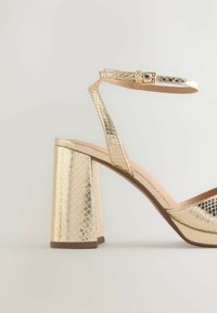 Sandale à talon haut dorée métallique avec talon bloc, bride à la cheville et finition en cuir texturé sur un fond clair uni.