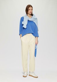 Un pull surdimensionné bleu avec des manches trois-quarts, un pantalon large crème et des baskets beige. Une écharpe en tricot bleu clair drape sur le pull.