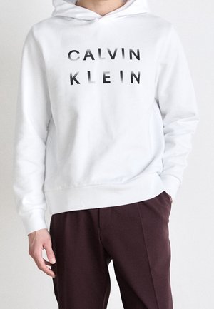 Persona que lleva una sudadera blanca con capucha con el texto "CALVIN KLEIN" en negro y pantalones marrón oscuro, de pie con una mano en el bolsillo.