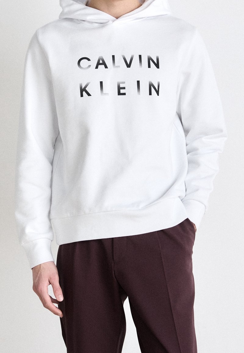 Personne portant un sweat à capuche blanc avec le texte « CALVIN KLEIN » en noir et un pantalon marron foncé, debout avec une main dans la poche.