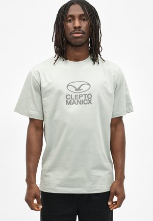 Mann mit langen Dreadlocks, der ein hellgraues T-Shirt mit dem "CLEPTO MANICX"-Logo und schwarze Hosen trägt, steht vor einem schlichten weißen Hintergrund.