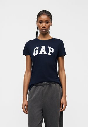 Jeune femme aux cheveux tressés portant un t-shirt GAP bleu marine et un pantalon gris foncé, debout sur un fond blanc uni.